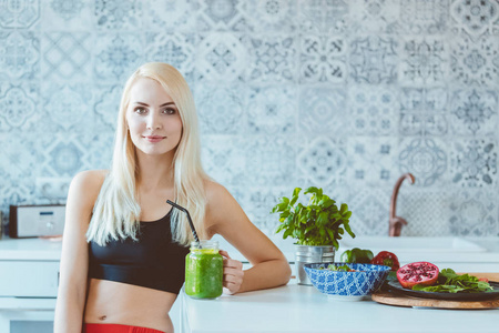 美丽的年轻女子穿着运动服，坐在厨房里，手里拿着绿色的冰沙，对着镜头微笑。
