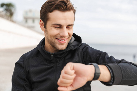 30多岁身穿黑色运动服的微笑男子坐在海边的木板路上看着手表的画面