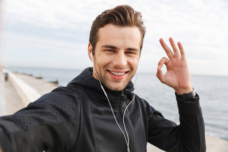 在海边散步时，身穿黑色运动服和耳机在手机上自拍照片的30多岁的运动员形象