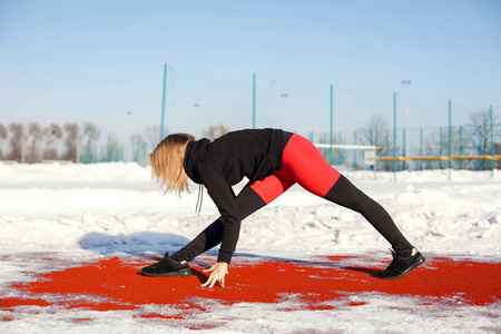 年轻的白种人女性金发碧眼，穿着紫罗兰色的紧身裤，在雪地体育场的红色跑道上伸展运动。 健康和运动生活方式。