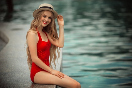 美丽的微笑的女孩穿着红色的泳衣, 靠在游泳池附近, 有蓝色的水。暑假