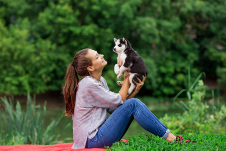 一个美丽的微笑的女人, 马尾和条纹衬衫抱着一个甜蜜的哈士基小狗, 而在草坪上的红色毯子上休息。对宠物的爱和关怀