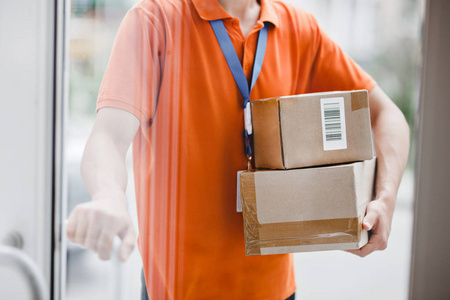 一名身穿橙色 t恤和名牌的人站在玻璃门后面, 拿着门把手和包裹给客户。友好的工人, 优质的送货服务