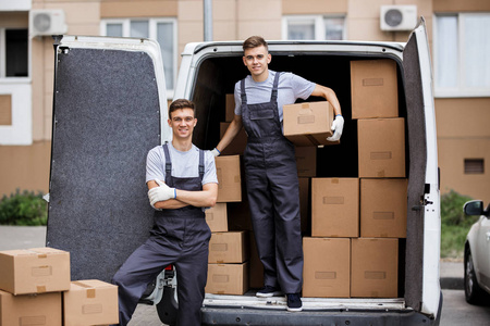 两个穿着制服的年轻英俊微笑的搬运工正在卸货, 面包车里装满了箱子。房子移动, 搬运工服务