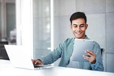 快乐的年轻亚洲商人在他的工作场所工作电脑笔记本电脑。微笑着看着数码平板电脑