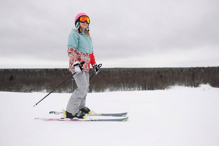 一个穿着极限运动服帽子和头盔戴着口罩的美丽的年轻咀嚼女子在森林的背景下滑雪