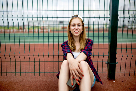 一个漂亮的微笑的金发女孩穿着格子衬衫, 白色帽子和牛仔短裤正坐在运动场上的护栏上, 手里拿着一部手机。运动和清凉风格