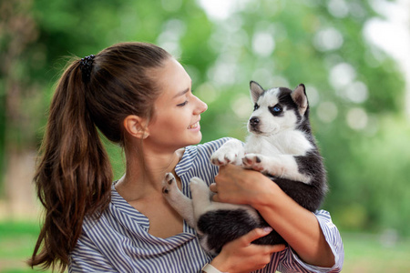 一个美丽的微笑的女人, 一条马尾, 穿着条纹衬衫, 站在草坪上抱着一只可爱的哈士奇小狗。对宠物的爱和关怀