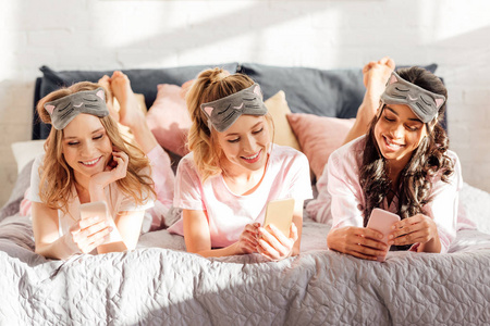 美丽的微笑的多元文化女孩，戴着睡面具躺在床上，在睡衣派对期间使用智能手机