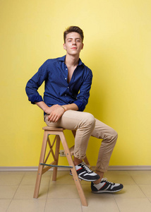 平静英俊的黑发男人穿着蓝色衬衫和米色裤子坐在黄色的墙壁上的木凳