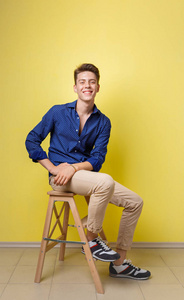 英俊友好的样子开朗的欧洲家伙穿着蓝色衬衫和米色裤子微笑, 而坐在黄色墙壁上的木凳