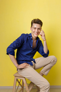 英俊友好的样子开朗的欧洲家伙穿着蓝色衬衫和米色裤子微笑, 而坐在黄色墙壁上的木凳