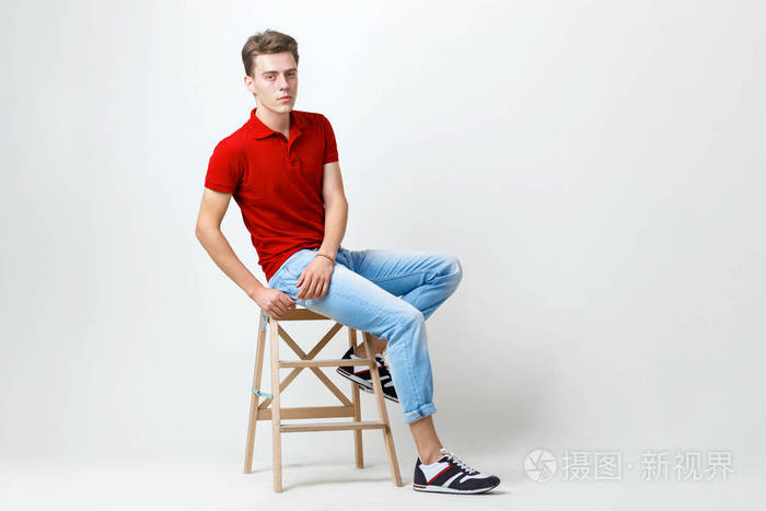 平静英俊的黑发男人穿着红色衬衫和蓝色牛仔裤坐在木制凳子上白色背景