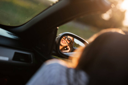 一个戴着太阳镜的微笑的年轻黑发女孩在汽车的侧镜中的倒影