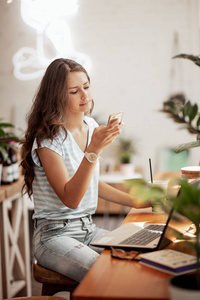 一个长头发的年轻苗条女孩, 穿着休闲的风格, 坐在桌旁, 拿着笔记本电脑, 在舒适的咖啡店里看着她的手机