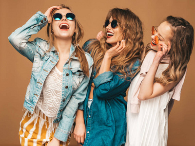 三个年轻美丽的微笑时髦女孩穿着时髦的夏季休闲牛仔裤衣服。 无忧无虑的女人在金色的墙壁附近摆姿势。 积极的模特在太阳镜上疯狂