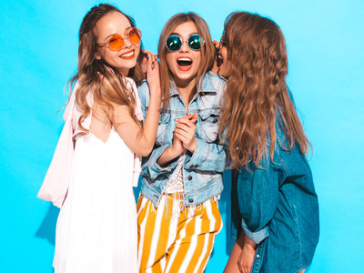 三个年轻美丽微笑的时髦女孩穿着时髦的夏季便服和圆形太阳镜。的女人分享秘密，八卦。孤立在蓝色上。令人惊讶的表情