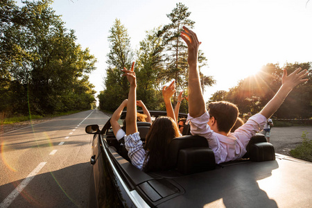 在一个温暖的阳光明媚的日子里, 年轻人乘坐敞篷车在路上举起手来.
