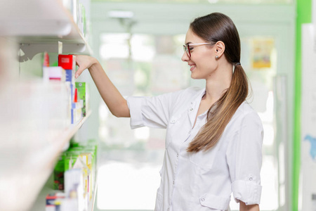 黑头发, 戴着眼镜, 穿着实验室的外套, 从现代药店的架子上拿东西