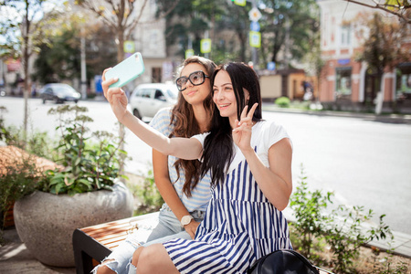 两个长着黑发的漂亮的瘦女孩, 穿着随意的衣服, 坐在板凳上自拍自拍