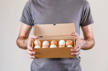 白人男子穿着灰色的t恤，在白色的背景上拿着一个装满母鸡蛋的纸板蛋盒。