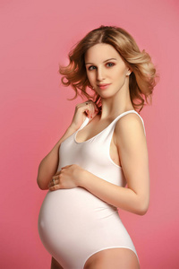 一位美丽的金发孕妇，穿着白色紧身衣，站在画室，穿着粉红色背景的女人微笑着