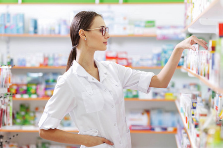 一个戴着眼镜的年轻的棕色头发的女孩, 穿着实验室的外套, 从药店的架子上拿了一些药。显示女孩的个人资料