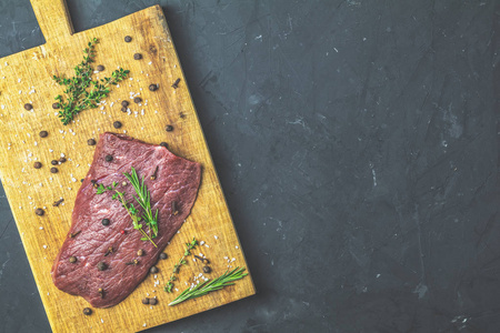 新鲜生肉牛肉牛排在木切割板上，配料为烹饪香料，百里香，迷迭香和橄榄油，深色背景，顶部视图复制空间为您提供文本。