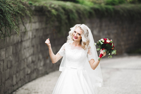 美丽的新娘在优雅的白色连衣裙，抱着花束摆在公园里