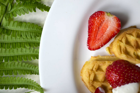 盘子里放着草莓的华夫饼，桌子上放着蕨类植物的绿叶。快关门。美味可口的早餐。