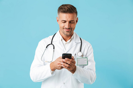 身穿白色外套手持智能手机站在蓝色背景上的英俊医生照片