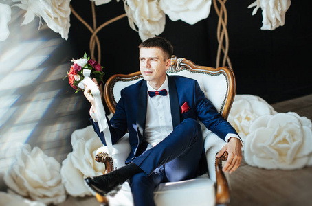 结婚西装新郎穿着时尚的蓝色西装,坐在一张白色的老式椅子上,在一朵