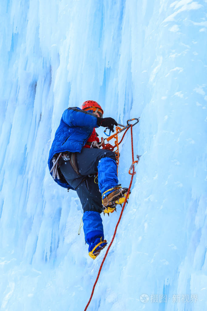 在下雪时攀岩者爬上了垂直的蓝色冰墙