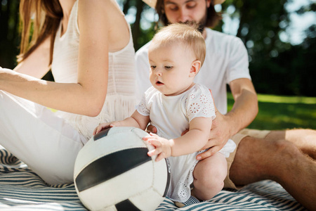 可爱的小女孩穿着白色礼服坐在父母旁边的毯子上的草坪上, 触摸足球