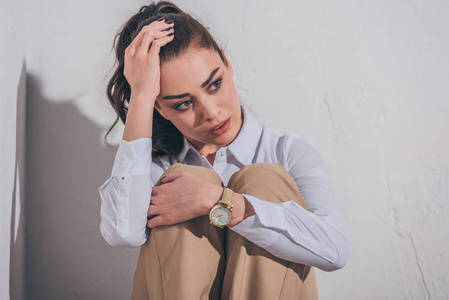 穿着衬衫和米色裤子的悲伤女人坐在房间的白色背景上悲伤混乱的概念