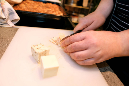 女人手切奶酪，她在厨房准备食物。厨师在餐厅餐桌上的木板上用刀切奶酪。家庭主妇拿着刀，切了块奶酪。