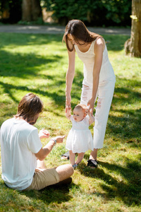 迷人的母亲和快乐的爸爸正在教他们的小女儿穿白色衣服如何迈出第一步