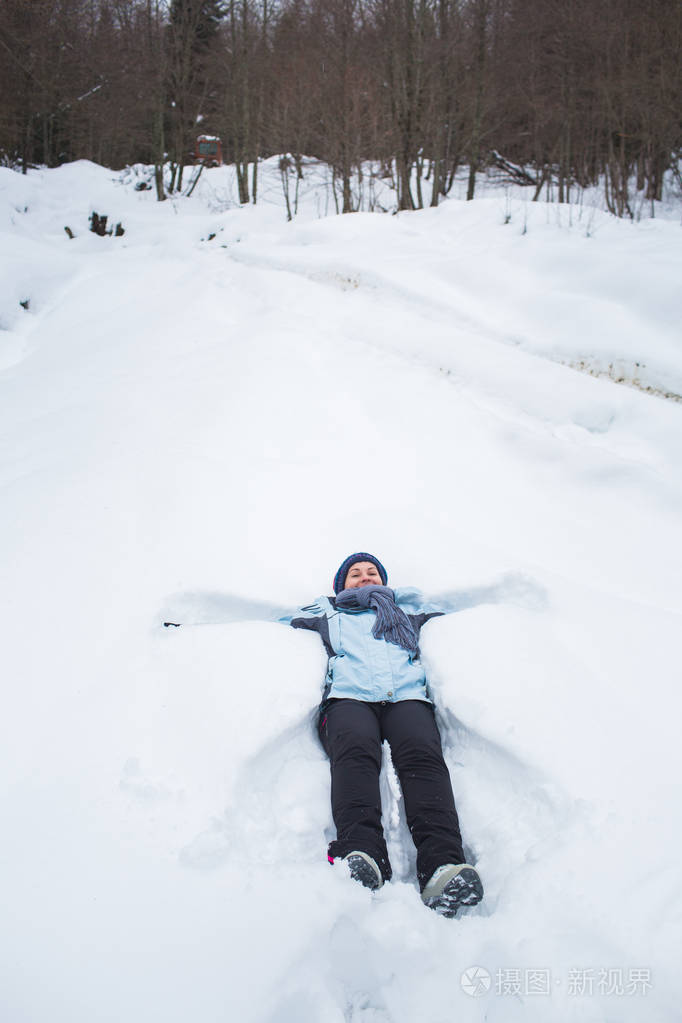 穿着裤子,夹克,手套和围巾的女孩躺在雪地里,在森林里堆雪天使