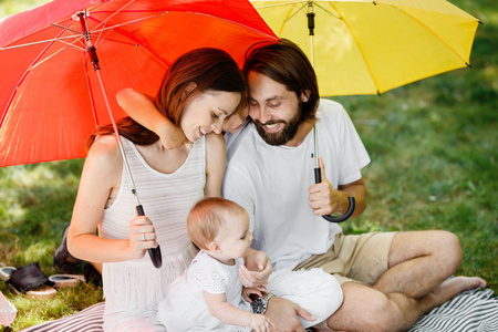 穿着白色衣服的时尚父母坐在红色和黄色雨伞下的毯子上, 小女儿