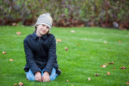 穿着黑色夹克和蓝色牛仔裤的微笑女孩坐在秋叶间的绿色草坪上