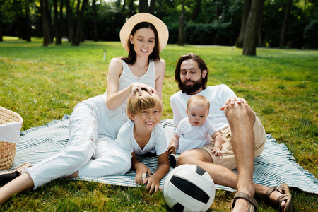 在公园里一个温暖阳光明媚的日子里, 穿着白色衣服的美丽的年轻家庭坐在草坪上的毯子上