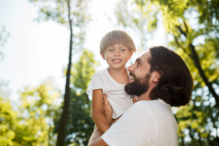 穿着白色 t恤的英俊的黑发父亲抱着他的小儿子, 看着他, 在公园里微笑着