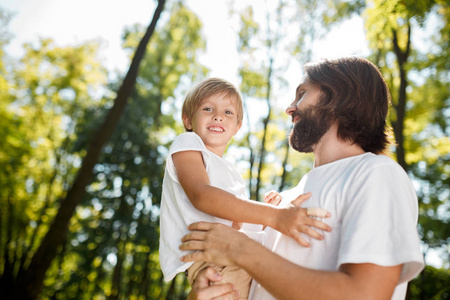 穿着白色 t恤的英俊的黑发父亲抱着他的小儿子, 在公园的一个夏天看着他