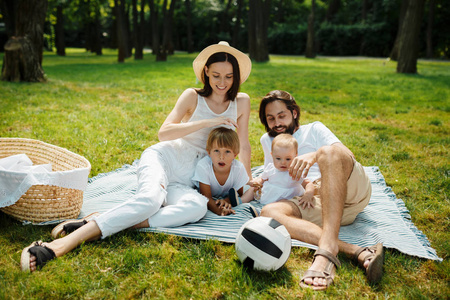 在公园里一个温暖阳光明媚的日子里, 穿着白色衣服的美丽的年轻家庭坐在草坪上的毯子上