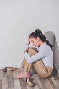 悲伤的女人穿着白色衬衫和米色裤子，坐在靠近墙壁的地板上，在家中悲伤混乱的概念