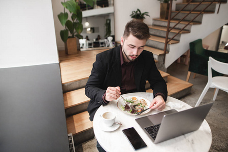 一个男人坐在一家舒适的餐厅里，桌子上放着一台笔记本电脑和一部智能手机，吃着沙拉。 忙碌的男人在咖啡馆里洗碗。
