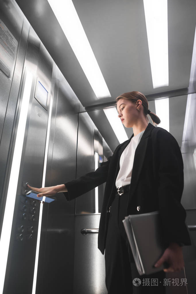 严肃的女商人按下电梯的按钮一只手拿着智能手机另一只手拿着笔记本