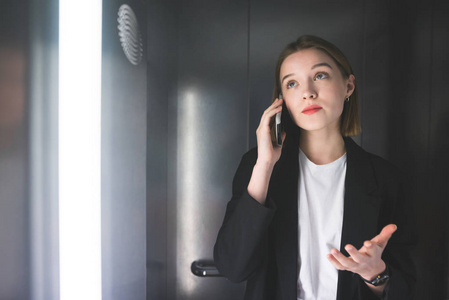 兴高采烈的女商人在电梯里用电话解释什么，穿着智能黑色西装。 年轻的女员工在她的智能手机上和她的老板和平交谈。