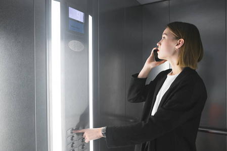 年轻的员工在电梯里用她的手机说话,按下楼层号码的按钮.照片