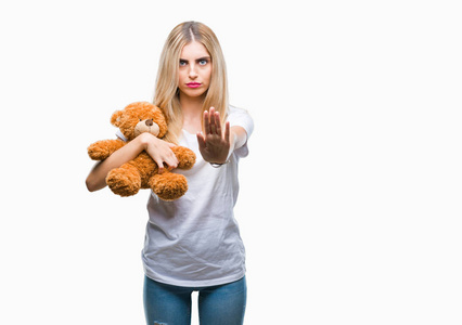年轻漂亮的金发女人，用张开的手在孤立的背景上抱着泰迪熊，做停止标志，严肃而自信的表情防御手势